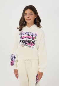 Ragazza che indossa una felpa crema e pantaloni con colorati testi e design in stile graffiti "Amiche per sempre" e "Ragazza fantastica".