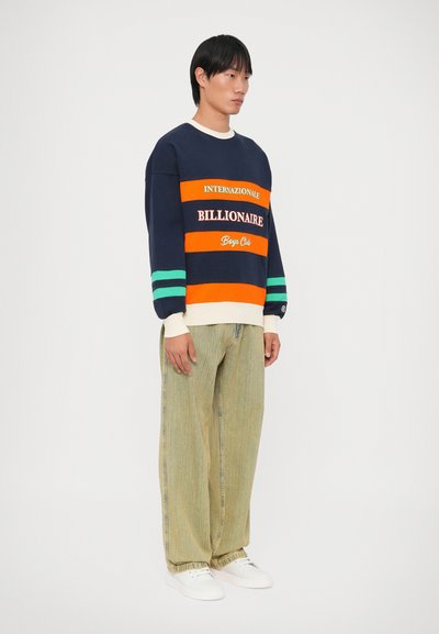 Sweatshirt azul-marinho com riscas laranja e verde, apresentando texto de logótype. Combinado com calças largas verde-claro e ténis brancos.