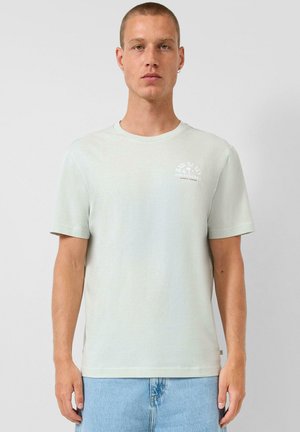 Młody mężczyzna z krótkimi włosami, ubrany w jasnooliwkową koszulkę i jasnoniebieskie dżinsy, stoi na tle jednolitego, szarego tła.