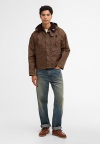 Brown waxed cotton jacket with a fur-lined collar, front pockets, and button closures, paired with blue jeans and brown leather shoes.