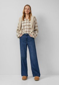 Frau steht mit den Händen in den Taschen, trägt ein kariertes Hemd, einen beigen Strickpullover über den Schultern, weit geschnittene Jeans und braune Schuhe vor einer schlichten Wand.
