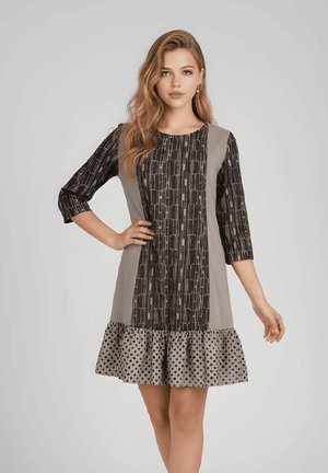 Joven mujer con cabello largo que lleva un vestido negro y gris con estampado, hasta la rodilla, con mangas tres cuartos y un dobladillo de lunares con volantes.
