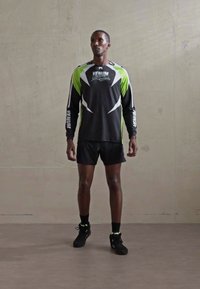 Mann steht in schwarzen Sportshorts, einem schwarzen und grünen langärmeligen Sporthemd mit "Venum"-Markierung und schwarzen Turnschuhen vor einer schlichten Wand.