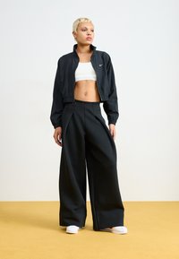 Sort cropped jakke med lynlås, hvid crop top og brede sorte bukser. Modellen har hvidt fodtøj på en gul baggrund.