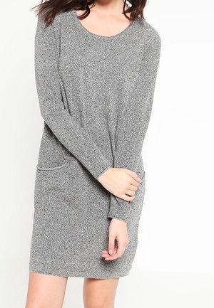 Femme portant une robe en maille grise à manches longues, longueur genou, avec un col rond et une petite poche sur le côté gauche.
