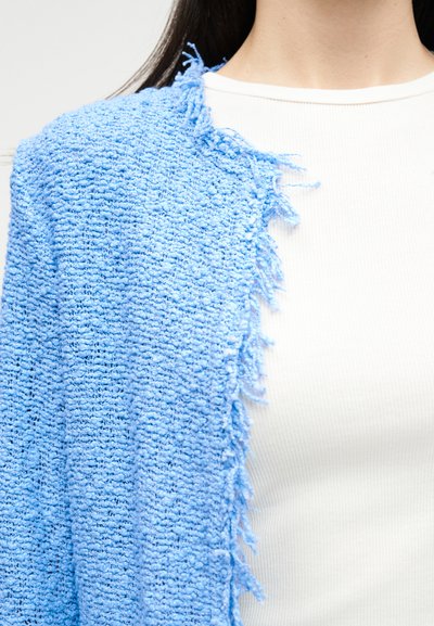 Άτομο που φοράει λευκό ριπ τοπ και υφασμάτινη ανοιχτό μπλε ζακέτα με ξεφτισμένες άκρες στον ώμο.