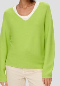 Grüner Strickpullover mit V-Ausschnitt, leichtem Material, langen Ärmeln und lässiger Passform, getragen über einem weißen schichteten Oberteil.