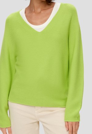 Pull en tricot vert avec un col en V, texture légère, manches longues et coupe décontractée, porté par-dessus un top blanc à plusieurs couches.