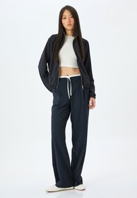 Marinblå zip-up jacka med elastiska ärmar och en kort vit topp, kombinerad med lösa marinblå byxor och vita sneakers, på en enkel bakgrund.