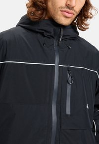 Zwarte jack met ritssluiting aan de voorkant en een capuchon, met reflecterende accenten en een borstzak. Gemaakt van lichte, gestructureerde stof.