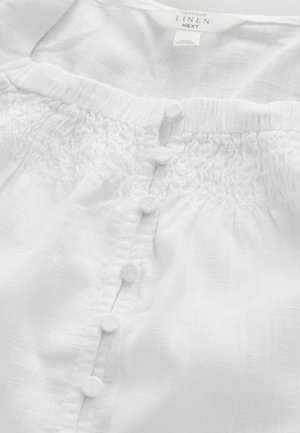 Vit linnesblus med rynkad halsringning och sex tygklädda knappar. Mjukt, strukturerat tyg med ett ljust, luftigt utseende.