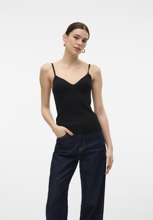Top nero a costine con scollatura a V e sottili spalline a spaghetti, abbinato a jeans in denim scuro a vita alta. La modella è in piedi con le mani nelle tasche.