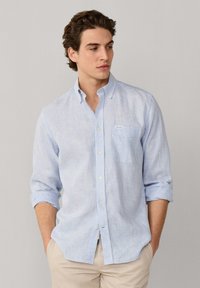 Hombre con una camisa de rayas blancas y azul claro de botones y mangas remangadas, pantalones beige, manos en los bolsillos, mirando hacia la izquierda.