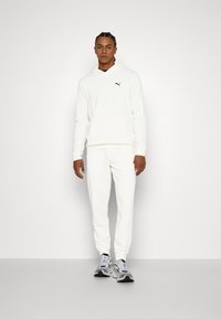 Hvid hoodie og matchende bukser, begge lavet af blødt materiale, med en kengurulomme og et lille sort logo. Sneakers i grå og sort.