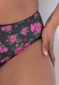 Zwarte bikinibroek met bloemenprint en roze rozen, voorzien van een gegolfde rand. De hand van het model rust op de stof, waarbij de rekbaarheid en textuur worden getoond.