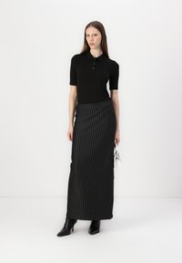 Černá polo košile s krátkými rukávy a knoflíkovou légu, v kombinaci s dlouhou sukní černo-bílého proužkovaného vzoru. Model má na sobě černé kotníkové boty.
