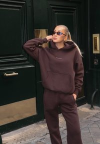 Brun fleece-hoodie med framficka och logotyp, matchad med träningsbyxor. Tillbehör inkluderar smala solglasögon och guldörhängen.