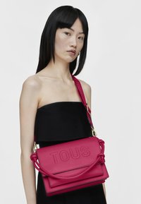Pink leather handbag with embossed "TOUS" logo, rectangular shape, detachable shoulder strap, and braided handle detail. Smooth texture.