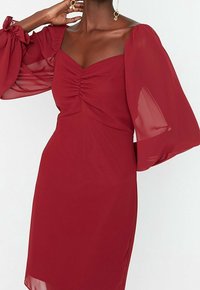 Rotes Kleid mit tailliertem Oberteil und gerafften Details, ausgestattet mit transparenten, puffigen langen Ärmeln und einem glatten, leichten Stoff.
