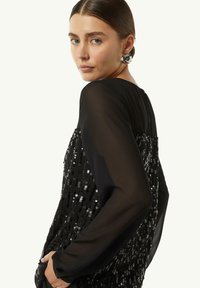 Schwarze Paillettenbluse mit durchsichtigen langen Ärmeln, Rundhalsausschnitt und zarter Textur. Die Pailletten erzeugen ein schimmerndes Muster über der Vorderseite.