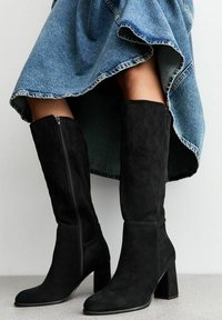 Stivali neri in suede alti fino al ginocchio con punta quadrata, tacco a blocco e cerniera laterale, abbinati a una gonna di denim con orlo sfrangiato.