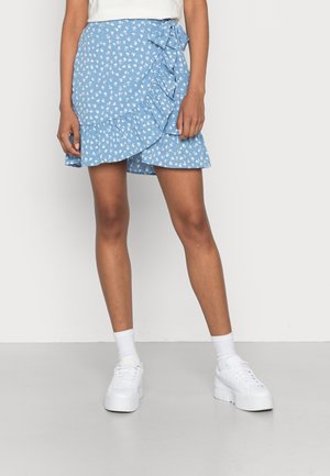 Femme portant une jupe portefeuille bleue à fleurs avec des volants, chaussettes blanches, baskets blanches à plateforme, debout les jambes légèrement écartées.