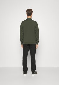 En man står vänd mot en vit vägg, klädd i en mörkgrön jacka, svarta byxor och svarta sneakers med vita sula.