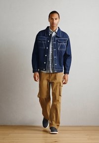 Veste en denim avec deux poches avant, superposée sur une chemise à carreaux. Pantalons cargo beiges avec des poches latérales, et baskets noires. Sol en bois clair.