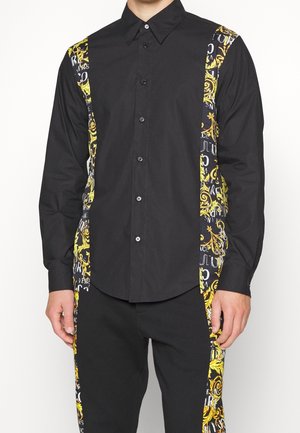 Camicia nera con bottoni con pannelli laterali decorati in giallo e bianco indossata da una persona in piedi.