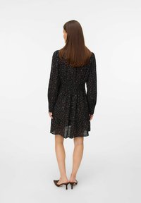 Femme aux longs cheveux bruns portant une robe noire à fleurs à manches longues et des mules à talons bruns, se tenant de dos sur un fond blanc uni.