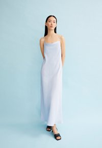 Robe maxi en satin bleu clair avec des fines bretelles, silhouette droite et texture lisse. Portée avec des sandales à talons noirs, sur un fond pâle.