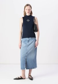 Marineblaues ärmelloses Oberteil mit Logo, kombiniert mit einem hellblauen Denim-Midirock mit Schlitz vorne. Schwarze Schultertasche und schwarze Ballerinas.