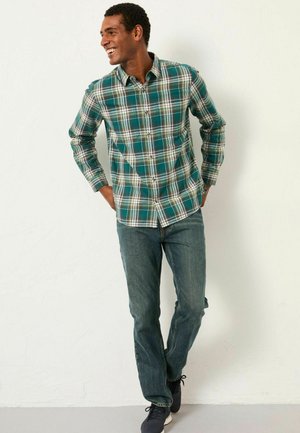 Grünes karierter Button-up-Hemd mit weißen und gelben Akzenten; kombiniert mit blauen Denim-Jeans und navyblauen Schuhen. Das Design hat lange Ärmel.