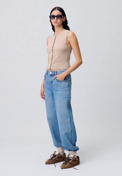 Top sin mangas de canalé beige con botones en el frente, combinado con unos vaqueros de pierna ancha de talle alto en azul claro y zapatillas marrones con cordones.