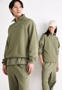Sudadera verde oliva con cremallera y logo bordado, combinada con pantalones de chándal a juego. Tejido suave, corte relajado y mangas con puños.