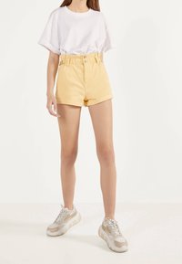 Gula denimshorts med hög midja och bältesöglor, kombinerade med en vit oversized T-shirt. Bärs med beigefärgade och grå sneakers.