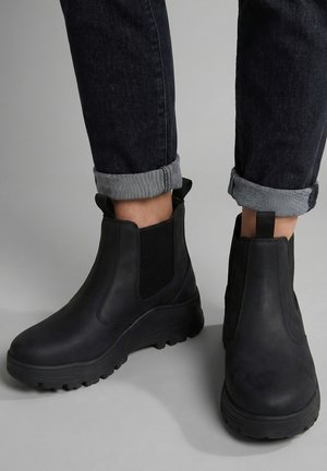 Persoon die zwarte leren Chelsea-laarzen draagt met dikke zolen en elastische zijpanelen, gecombineerd met opgerolde donkere jeans.