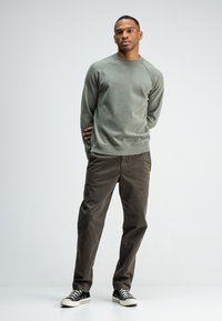 Grau-grüner Rundhalsausschnitt-Sweatshirt mit gerippten Bündchen, kombiniert mit braunen Chinos und schwarzen Sneakers, vor einem schlichten weißen Hintergrund.