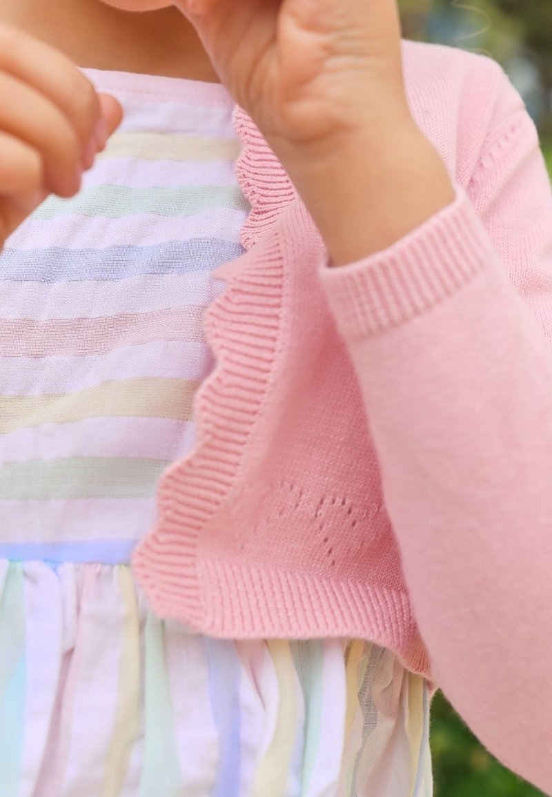 Jeune enfant portant une robe pastel rayée et un cardigan rose, tenant ses mains près du visage en plein air avec un arrière-plan flou.