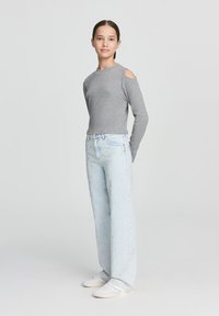 Maglietta grigia a maniche lunghe con aperture sulle spalle, abbinata a pantaloni blu chiaro a gamba larga con borchie decorative. Sneakers bianche completano il look.