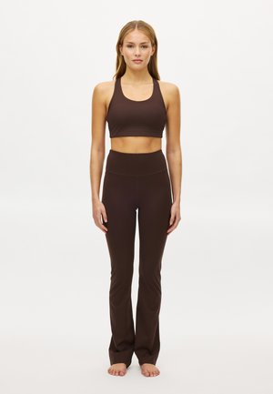 Kvinde stående barfodet iført en brun sports-bh og brune, højtaljede, udvidede leggings mod en ensfarvet hvid baggrund.