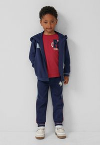 Marineblauwe hoodie en trainingsbroek set met een rood T-shirt, met contrasterende stiksels, sportief ontwerp en logodetails. Witte sneakers maken de look compleet.