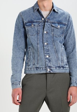 Mann trägt eine hellblaue Jeansjacke mit weißen Knöpfen und dunkelgraue Hose, steht vor einem einfarbigen Hintergrund.