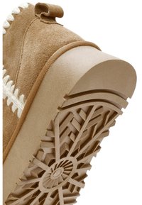 Bruine suède instap-schoen met een gestructureerde beige rubberen zool die een geometrisch profiel heeft en een kleine hak. Witte stiksels accentueren het ontwerp.