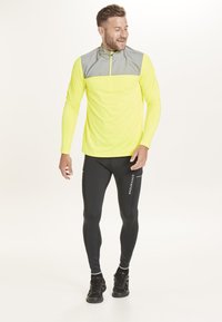 Langærmet atletisk top i neongul med grå detaljer, halv-zip krave; parret med sorte tætsiddende leggings med hvid tekst og detaljer.