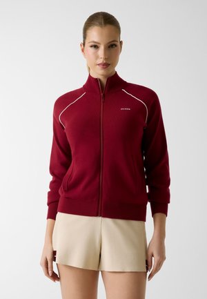 Femme portant un pull zippé bordeaux avec des passepoils blancs et un short beige, debout devant un fond clair uni.