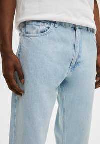 Jeans de mezclilla azul claro con un corte relajado, diseño de cinco bolsillos y detalles de botones de metal en la cintura. Se aprecia una textura desgastada.