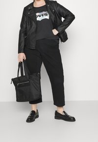 Czarna skórzana kurtka motocyklowa na czarnej koszulce z logo Levi's, w połączeniu z czarnymi krótkimi spodniami i błyszczącymi czarnymi mokasynami. Trzyma czarną torbę tote.