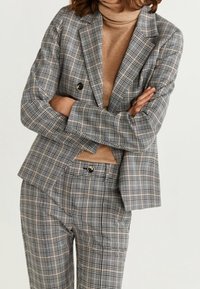 Šedý kostkovaný blazer a odpovídající kalhoty, se vzorem v béžové a černé barvě. Texturovaná látka se dvěma knoflíky na blazoru.