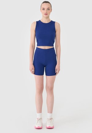 Femme debout, bras détendus, portant un crop top bleu sans manches et un short taille haute assorti, avec des chaussettes blanches et des baskets rose clair.